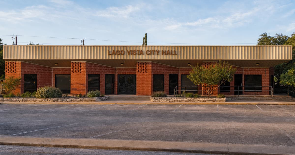 Lago Vista City Hall, a one-story red brick building with a tan metal canopy, in early morning light with an empty parking lot in front