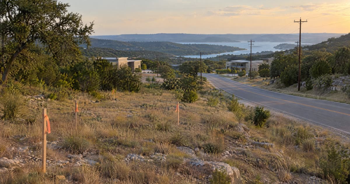 Survey stakes with orange flagging tape in a vacant, grassy lot beside a two-lane road, with low commercial buildings and Lake Travis in the distance at sunset